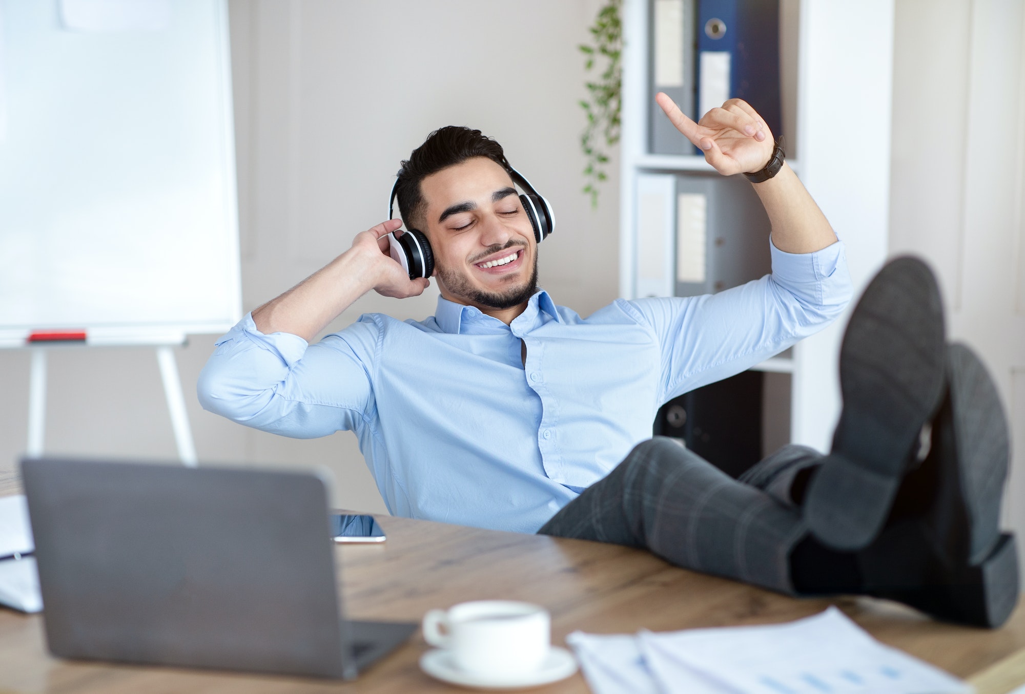 Taking break at office. Joyful Arab man listening to music in wireless headphones, relaxing at
